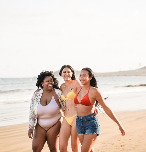 Drei junge Frauen mit unterschiedlichen Körperproportionen gehen zusammen in farbigen Bikinis am Strand entlang